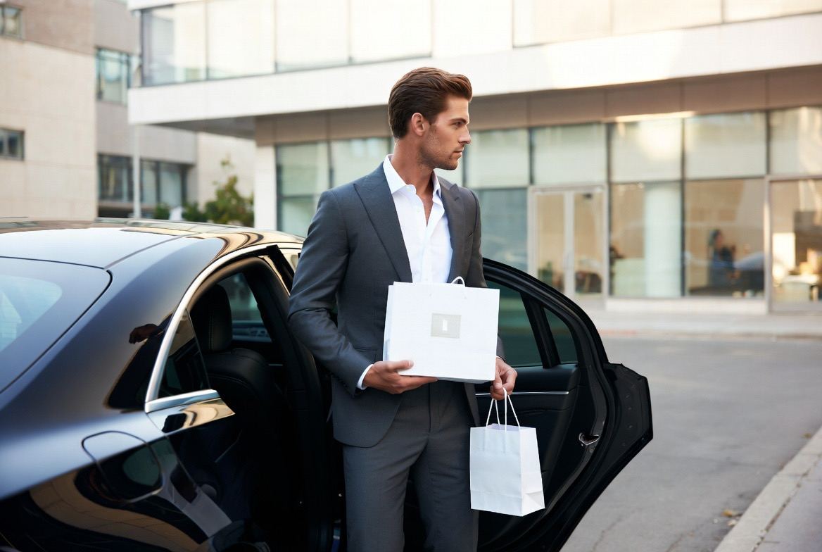 Concierge in a grey suit carrying a shopping bag and a boxed item to the open rear door of a black sedan.