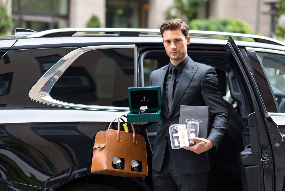 Concierge in a charcoal suit beside an open black SUV holding a presentation watch box, a leather two-bottle wine carrier and a sealed retail package.